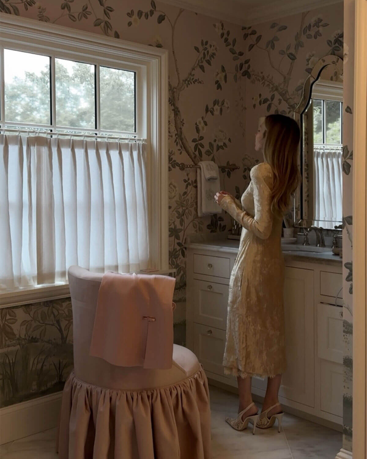 Emily wearing dress getting ready with vanity chair and floral wallpaper for bathroom